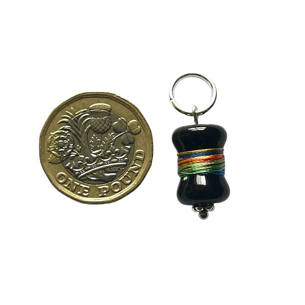 Black bobbin glass bead wrapped with multicolour thread handing from a jump ring to create a stitch marker. Shown with a pound coin for scale.