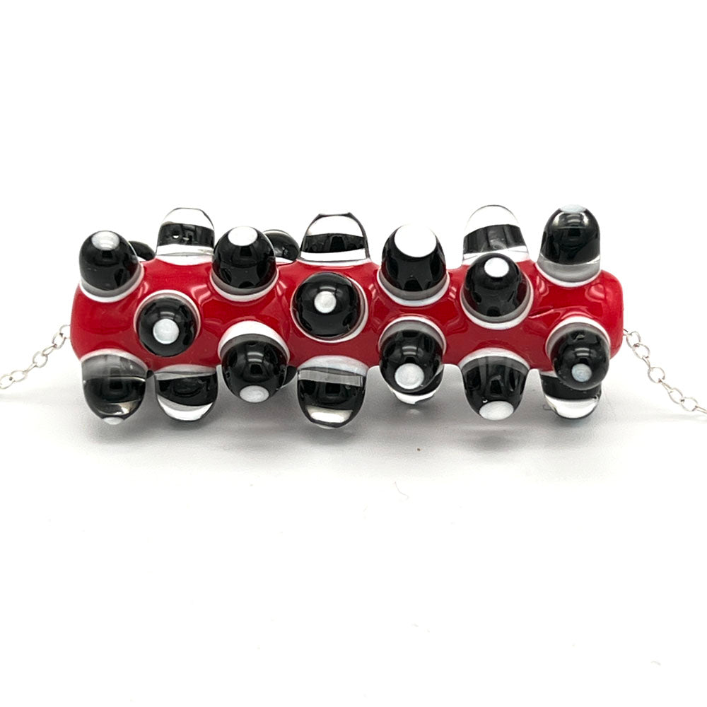 Close up of glass bead necklace. The bead is a long red tube which has been heavily decorated with dots of black, clear and white glass. The necklace is completed with a sterling silver chain.