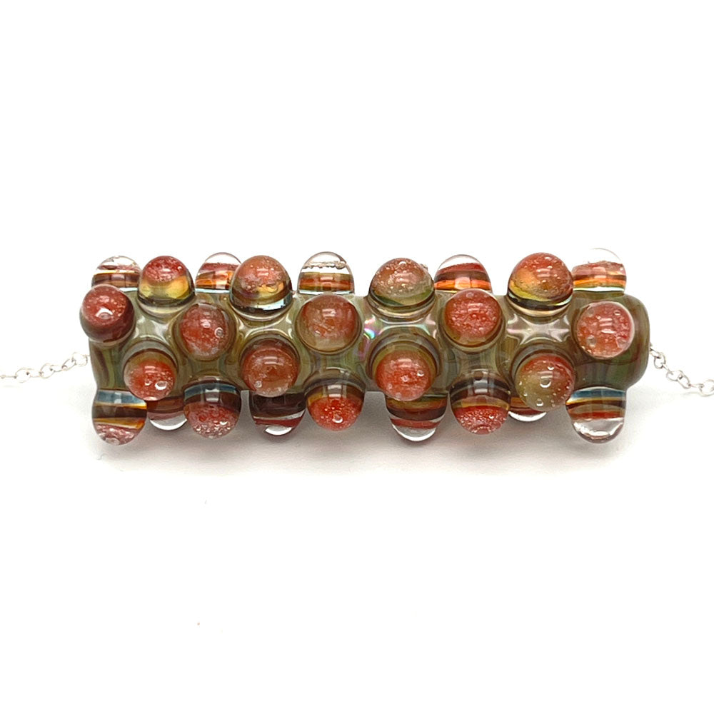 Close upof blossom bead necklace. The bead is a long sage green tube which is heavily decorated with raised dots. The dots are made of coloured layers of blue, yellow and red, all separated with clear glass. The necklace is finished with a sterling silver chain.