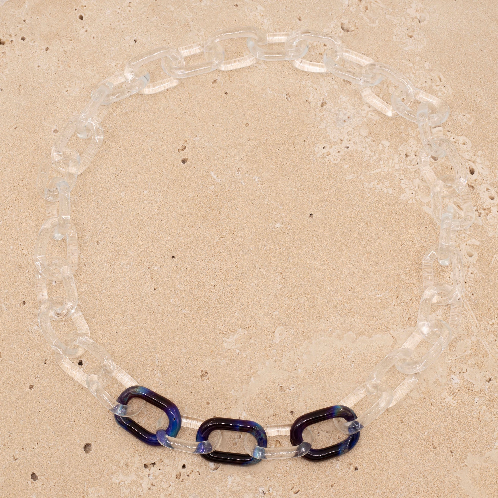 Overhead view, sandstone background, clear glass necklace with three blue links