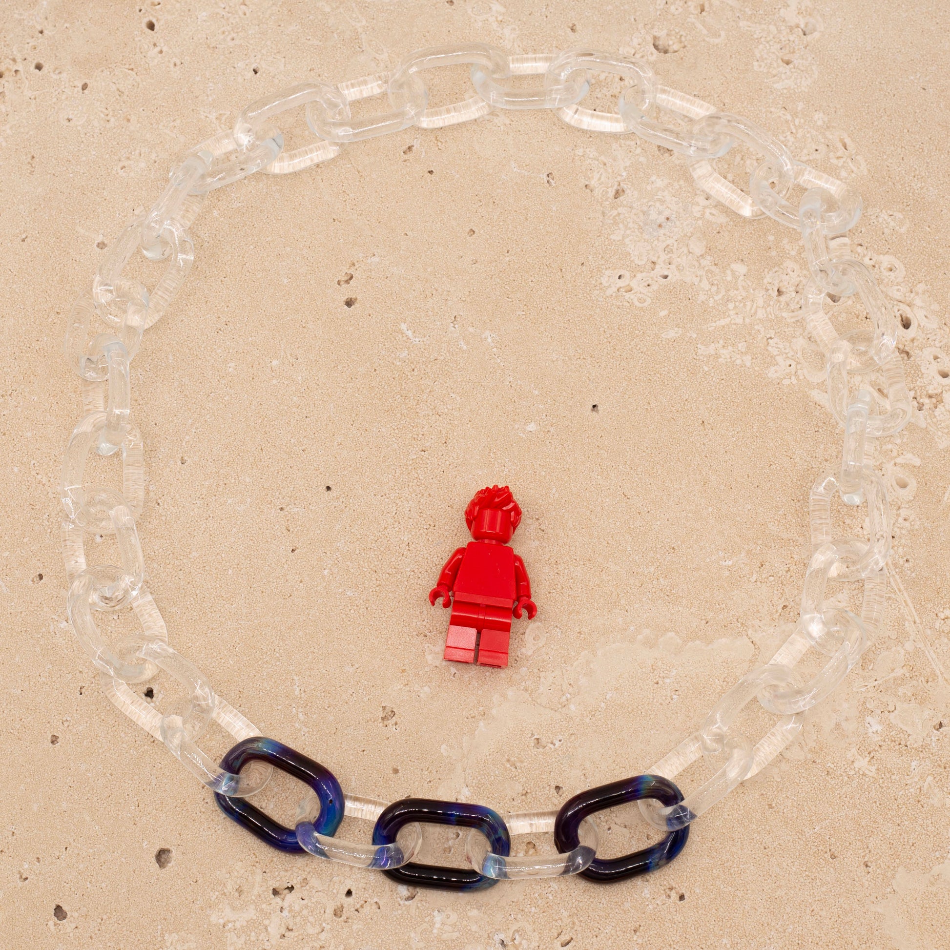 Overhead view of clear glass necklace with three blue links, sandstone background. Image includes Lego figure for scale
