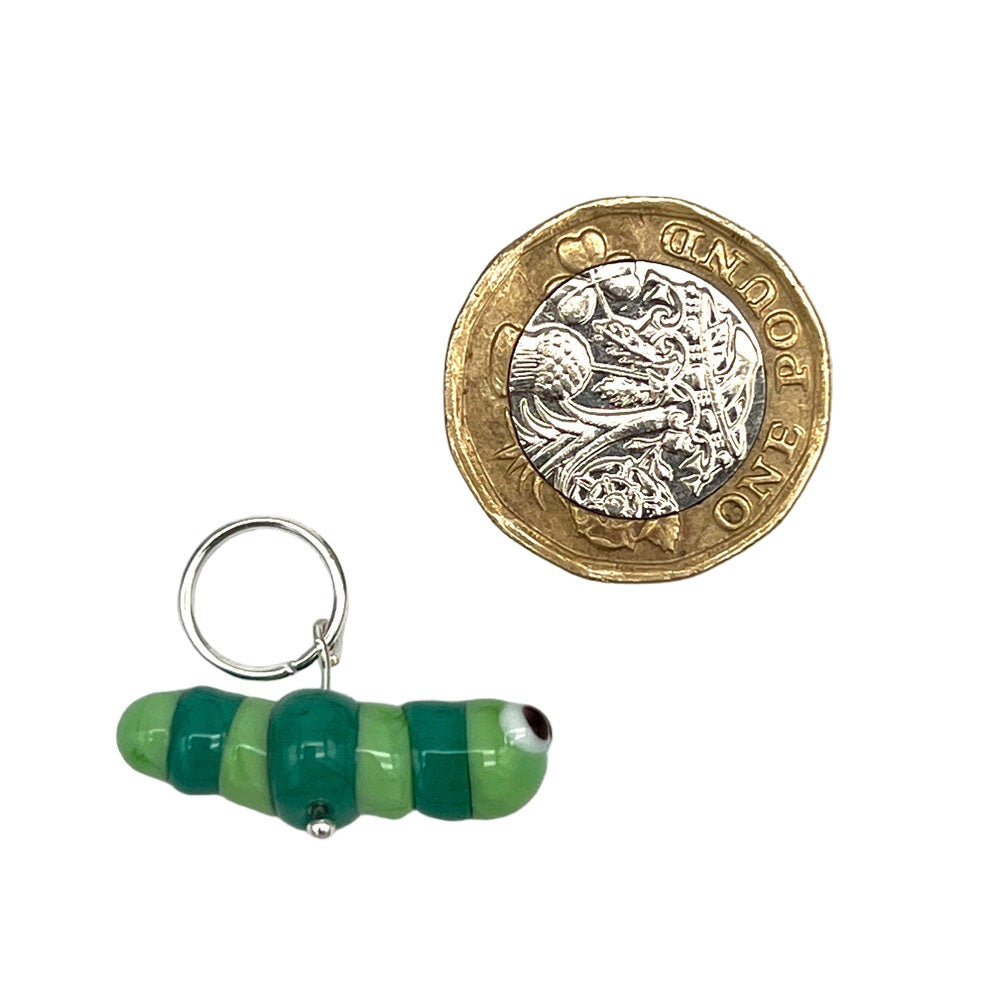 Close-up of a green and teal glass caterpillar stitch marker with a silver jump ring next to a one-pound coin for size comparison