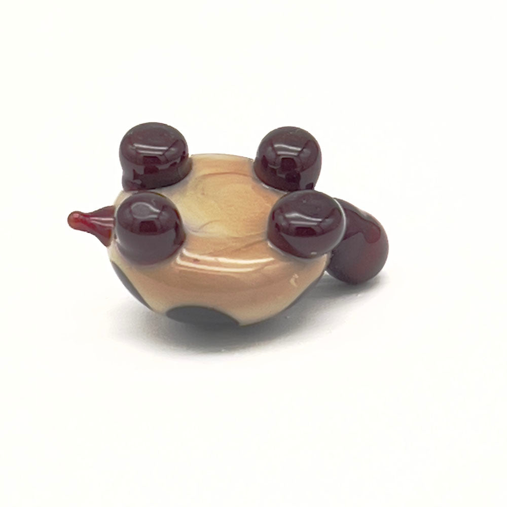 The underside of the same glass tortoise, showing its beige shell with dark brown spots and dark brown legs and head, laying upside down on a white surface.