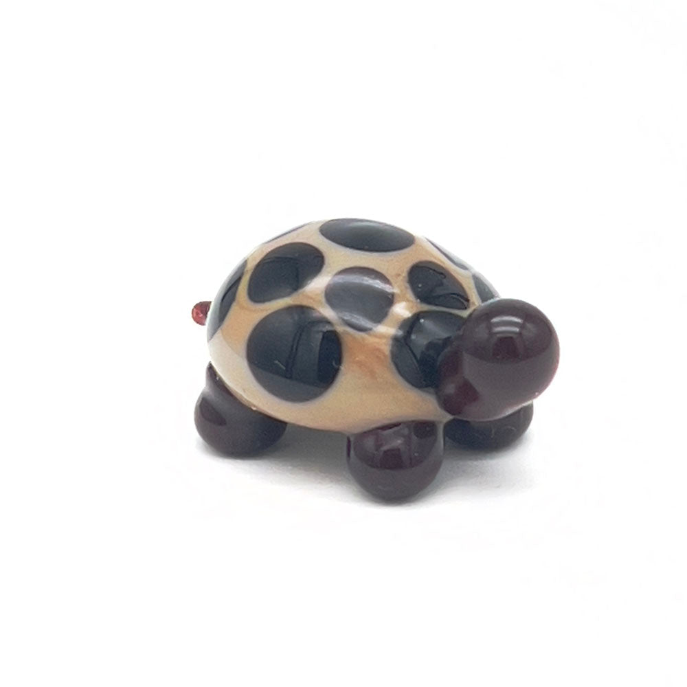 The front view of the glass tortoise, highlighting its beige shell with dark brown spots, dark brown head, and legs, positioned on a white background.