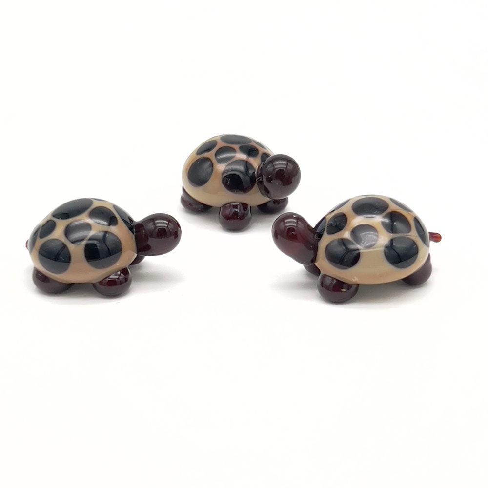 A group of three similar glass tortoises, each with a beige shell and dark brown spots, dark brown heads, and legs, arranged on a white background.