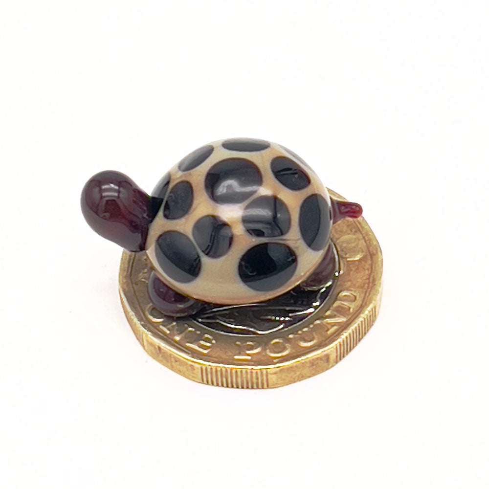The miniature glass tortoise sitting on a one-pound coin, highlighting its small size. The tortoise has a beige shell with dark brown spots, and dark brown head and legs.