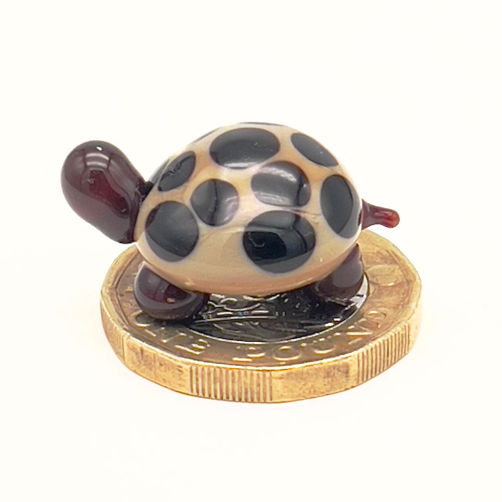 The glass tortoise on top of a one-pound coin, viewed from a different angle, emphasizing its small size. The tortoise features a beige shell with dark brown spots and dark brown head and legs.