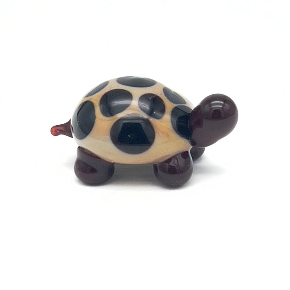 The side view of the glass tortoise, showcasing its beige shell with dark brown spots, dark brown head, and legs, displayed on a white background.