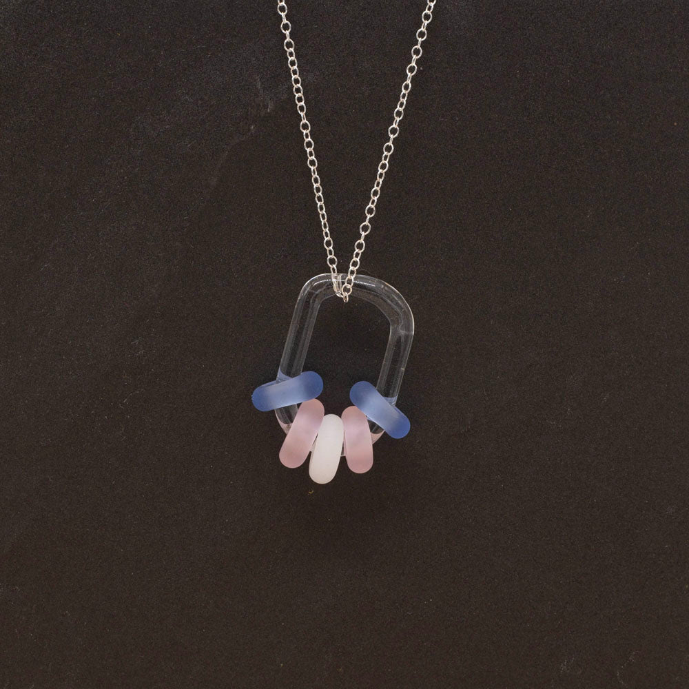 Clear glass link passing through blue, pink and white beads, hanging on a sterling silver chain, dark background.