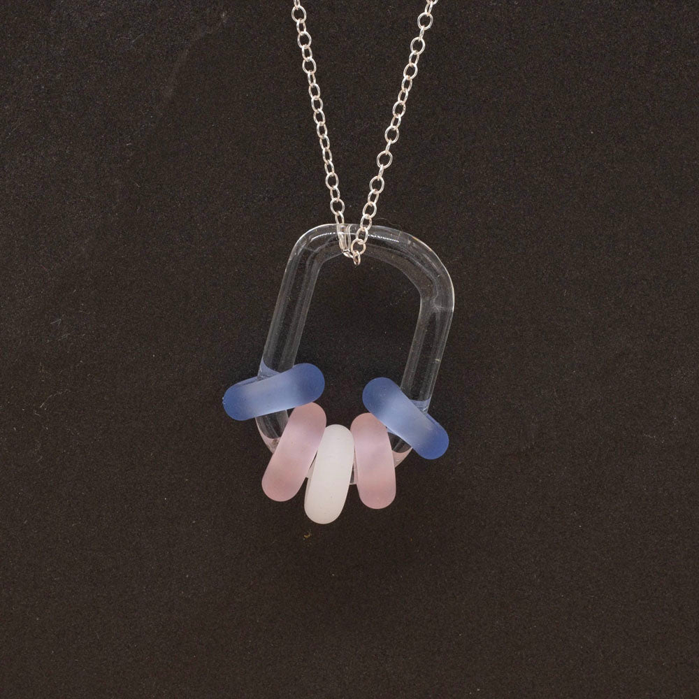 Close up of clear glass link passing through blue, pink and white beads, hanging on a sterling silver chain, dark background.
