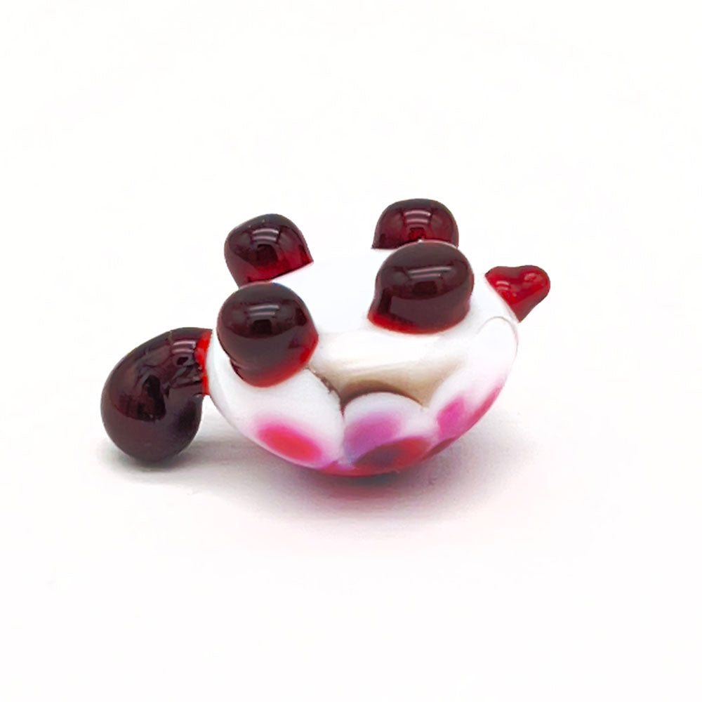 The underside of the same glass tortoise, showing its white shell with multicolored speckles and dark brown legs and head, laying upside down on a white surface.