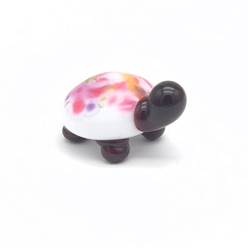 The front view of the glass tortoise, highlighting its white shell with multicolored speckles, dark brown head, and legs, positioned on a white background.