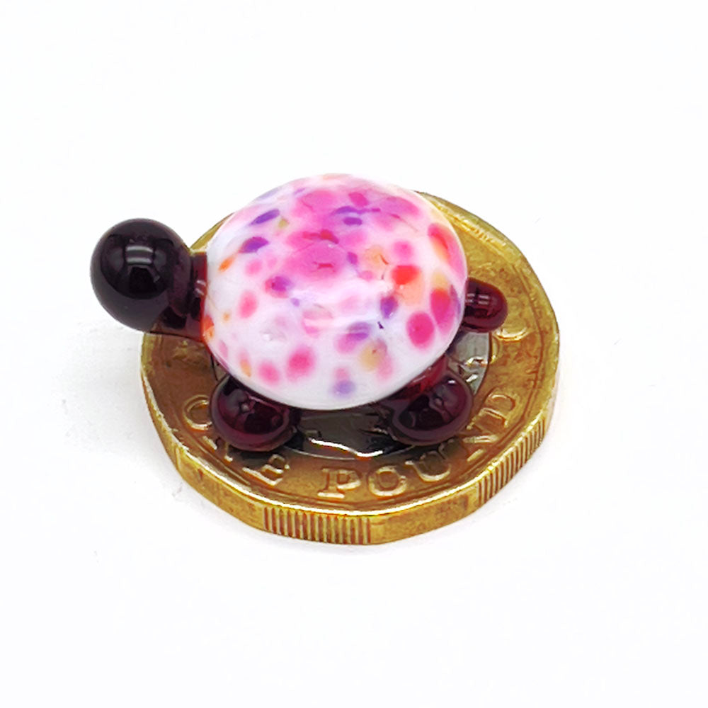 The miniature glass tortoise sitting on a one-pound coin, highlighting its small size. The tortoise has a white shell with multicolored speckles, and dark brown head and legs.