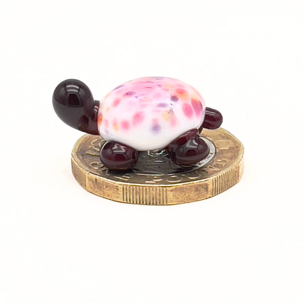 The glass tortoise on top of a one-pound coin, viewed from a different angle, emphasizing its small size. The tortoise features a white shell with multicolored speckles and dark brown head and legs.