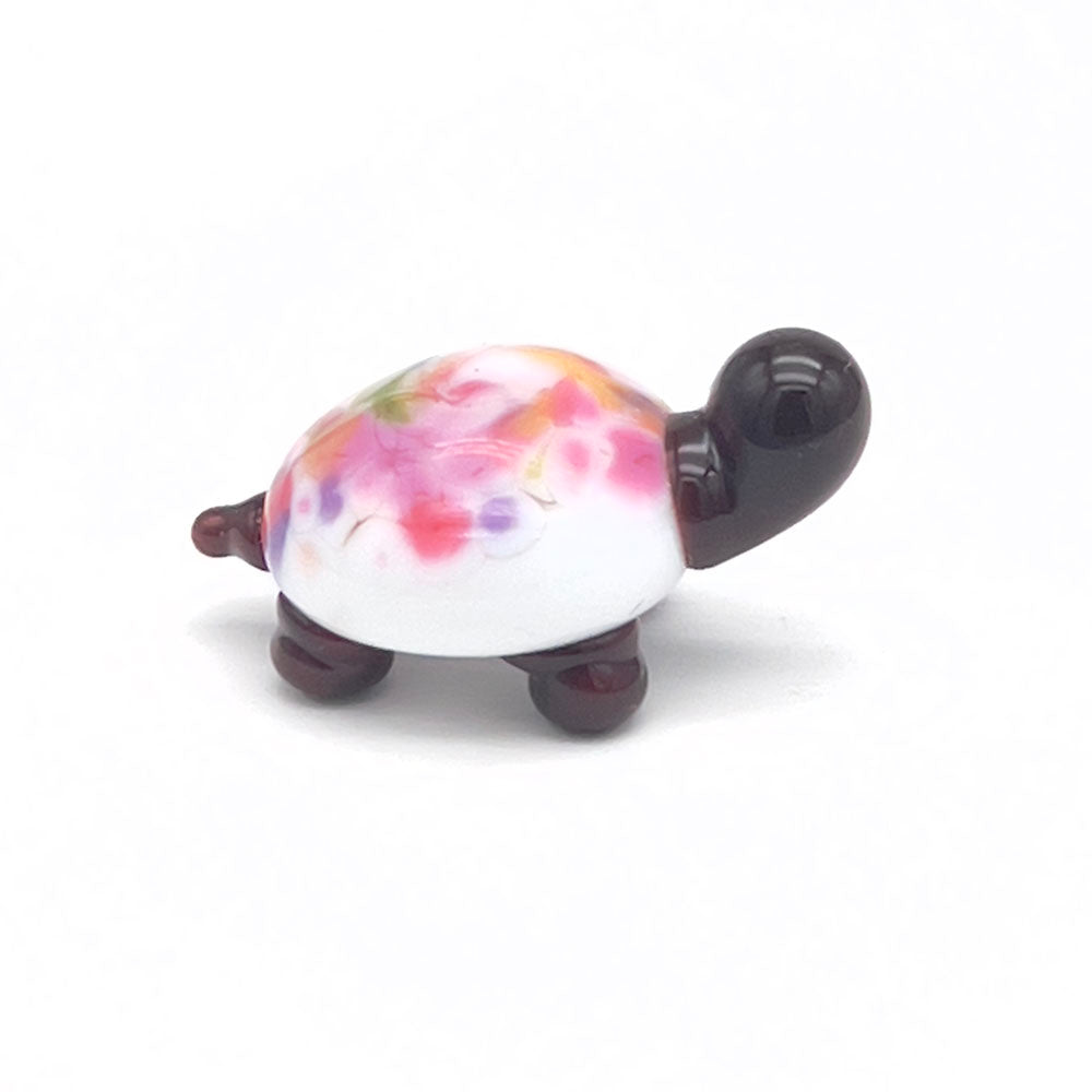 The side view of the glass tortoise, showcasing its white shell with multicolored speckles, dark brown head, and legs, displayed on a white background.