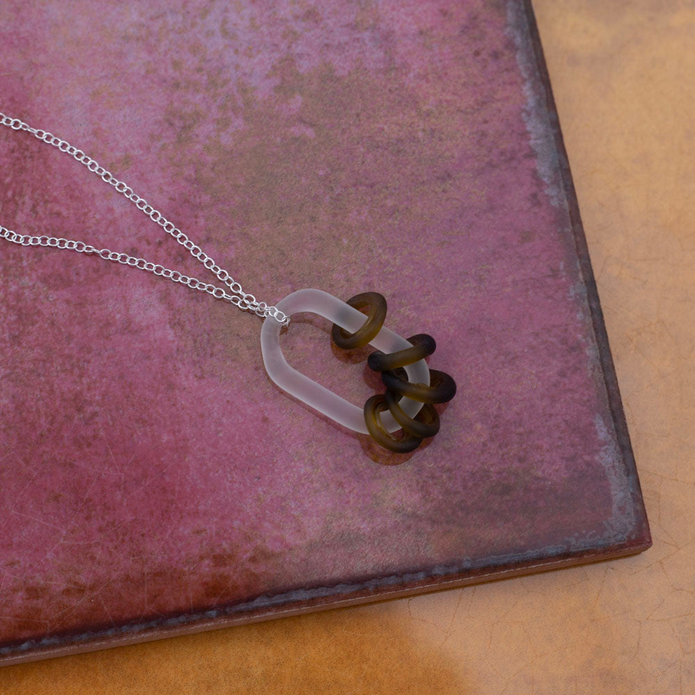 Photographed on pink and yellow tiles. A frosted link made from clear glass which passes through five beads. The beads are made from a marmite bottle so are brown. They also have a frosted finish. The link hangs from a sterling silver chain.