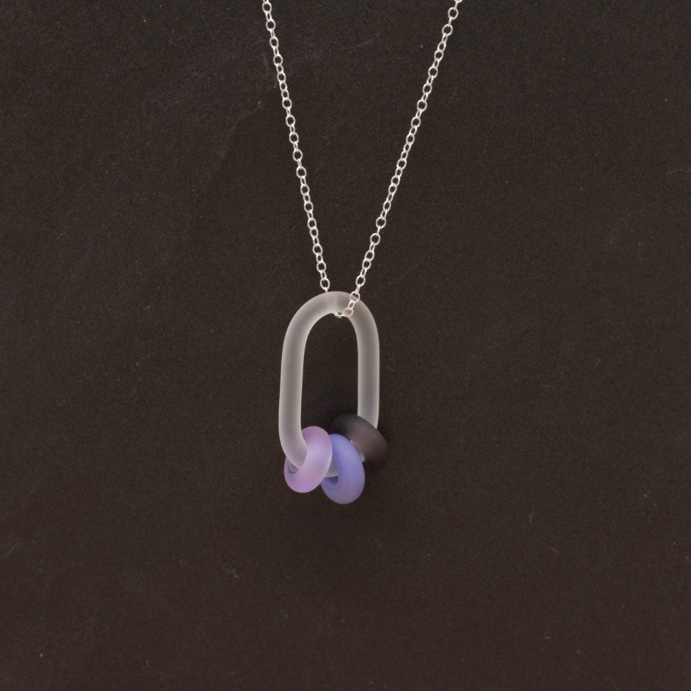 Clear link which passes through beads in three shades of purple glass. Link and beads have a frosted finish. The link hangs from a sterling silver chain. Shown on a dark background.