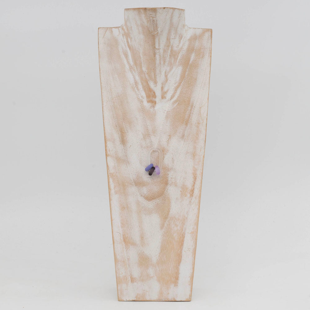 Clear link which passes through beads in three shades of purple glass. Link and beads have a frosted finish. The link hangs from a sterling silver chain. Shown on a whitewashed wooden bust.