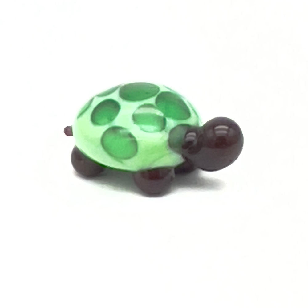 The front view of the glass tortoise, highlighting its light green shell with dark green spots, dark brown head, and legs, positioned on a white background.