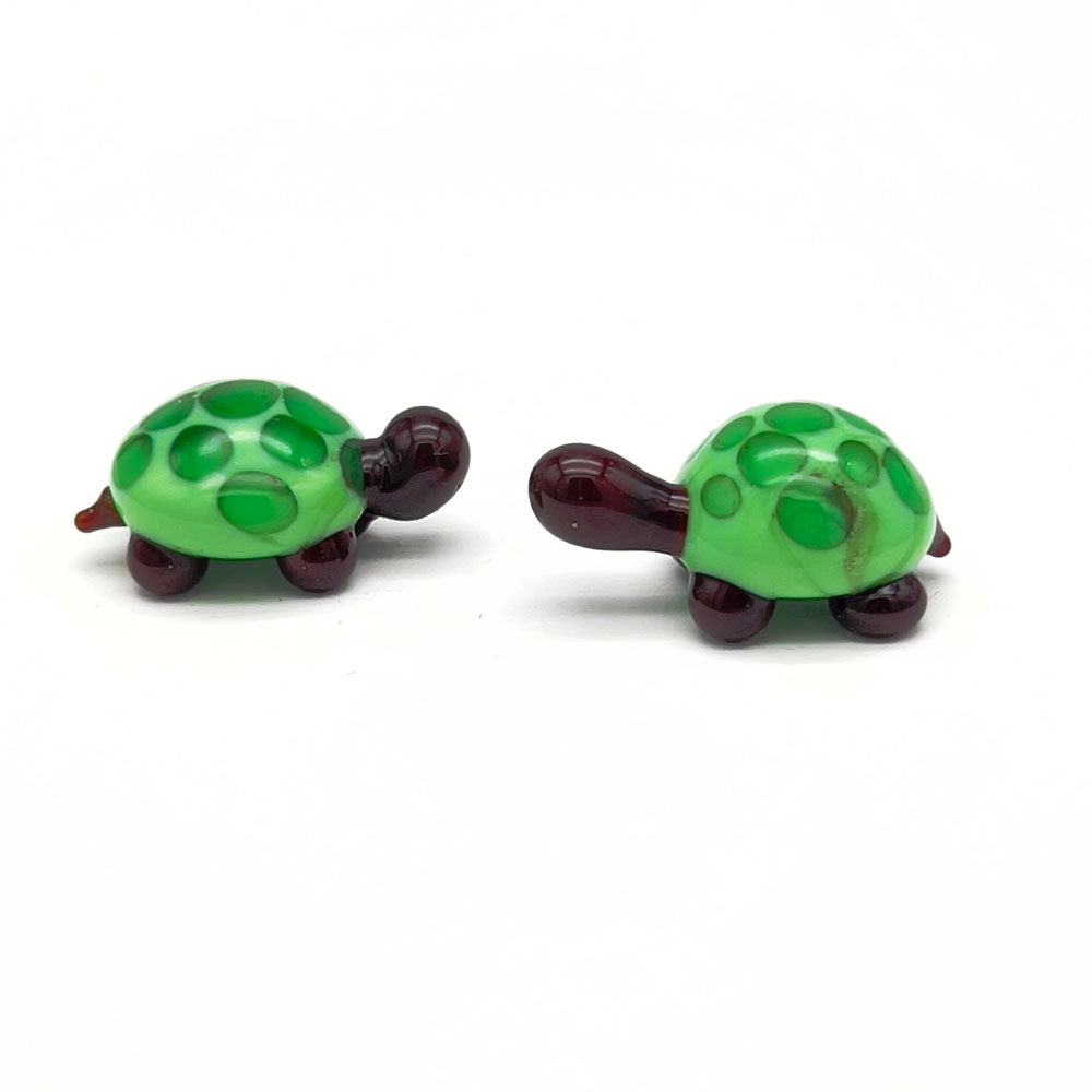 A pair of matching glass tortoises, each with a light green shell and dark green spots, dark brown heads, and legs, arranged on a white background.