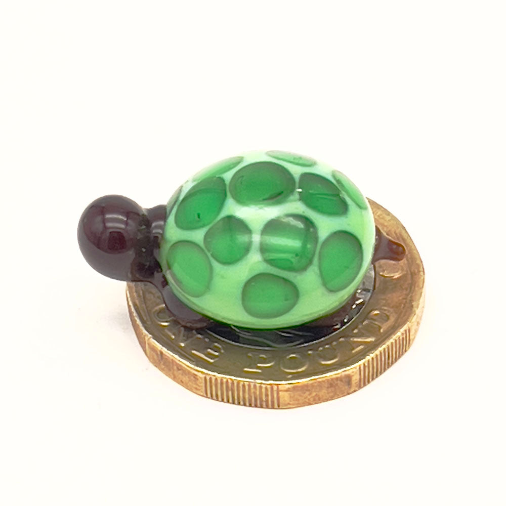 The miniature glass tortoise sitting on a one-pound coin, highlighting its small size. The tortoise has a light green shell with dark green spots, and dark brown head and legs.