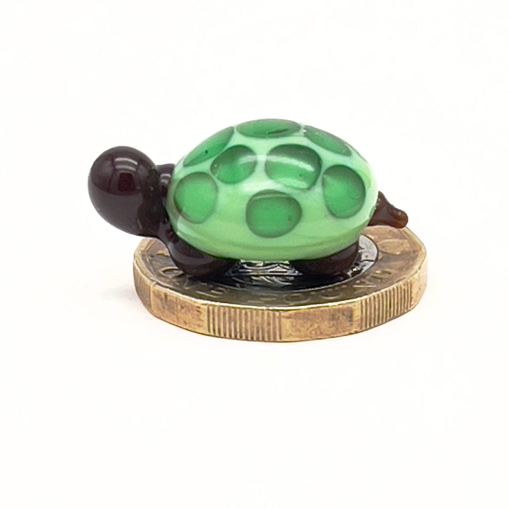The glass tortoise on top of a one-pound coin, viewed from a different angle, emphasizing its small size. The tortoise features a light green shell with dark green spots and dark brown head and legs.