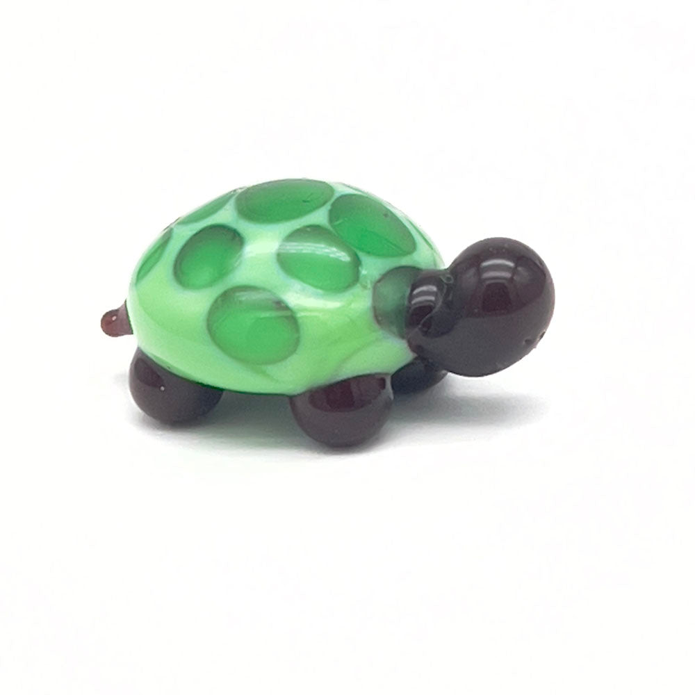 The side view of the glass tortoise, showcasing its light green shell with dark green spots, dark brown head, and legs, displayed on a white background.