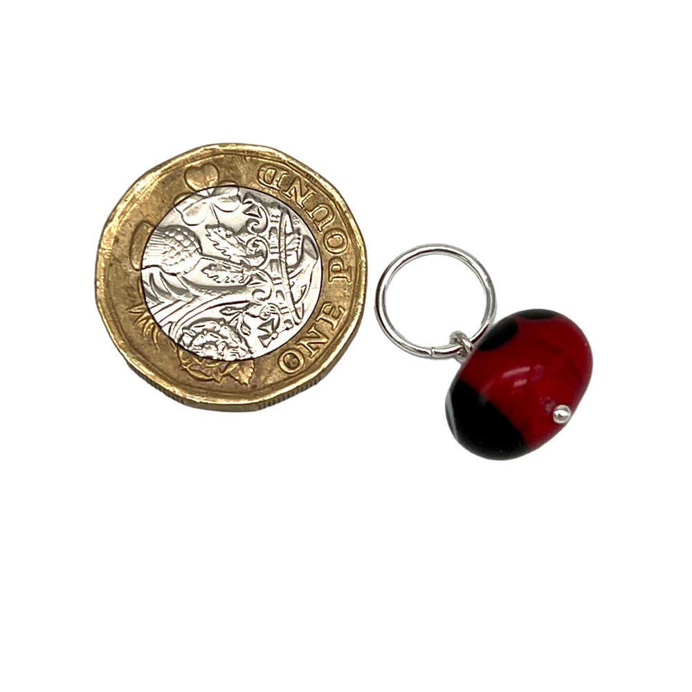 A single glass ladybird stitch marker with a jump ring, placed next to a pound coin for scale. The ladybird is red with black spots and details.