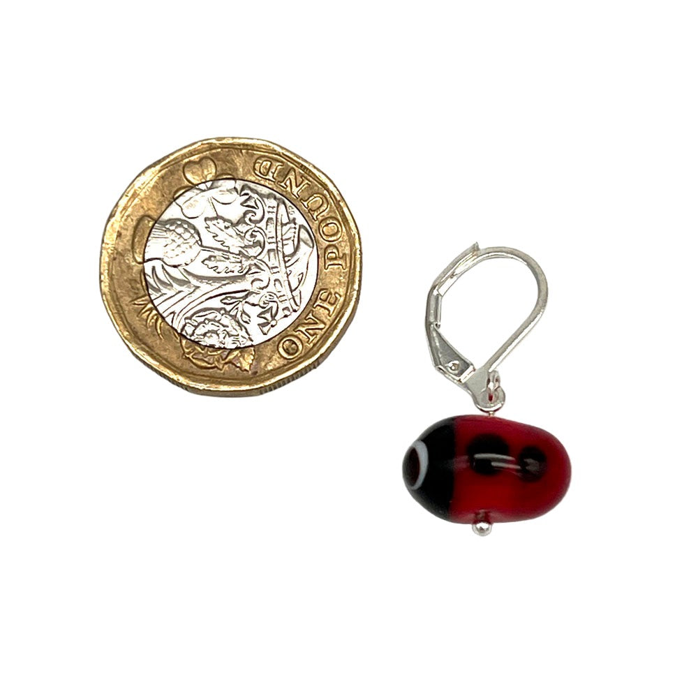 A single glass ladybird stitch marker with a latch back hook, placed next to a pound coin for scale. The ladybird is red with black spots and details.