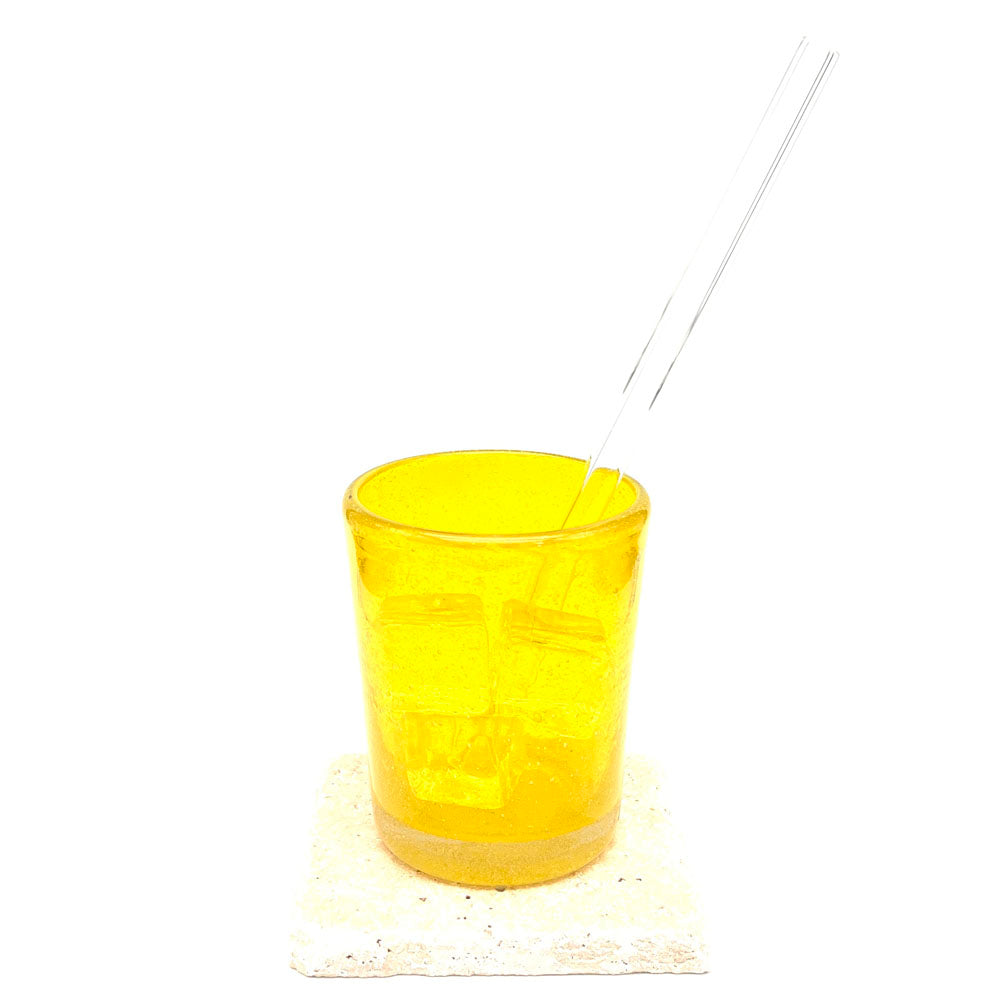 A long clear glass straw sits in a yellow tumbler filled with ice. The tumbler sits on a sand stone tile.