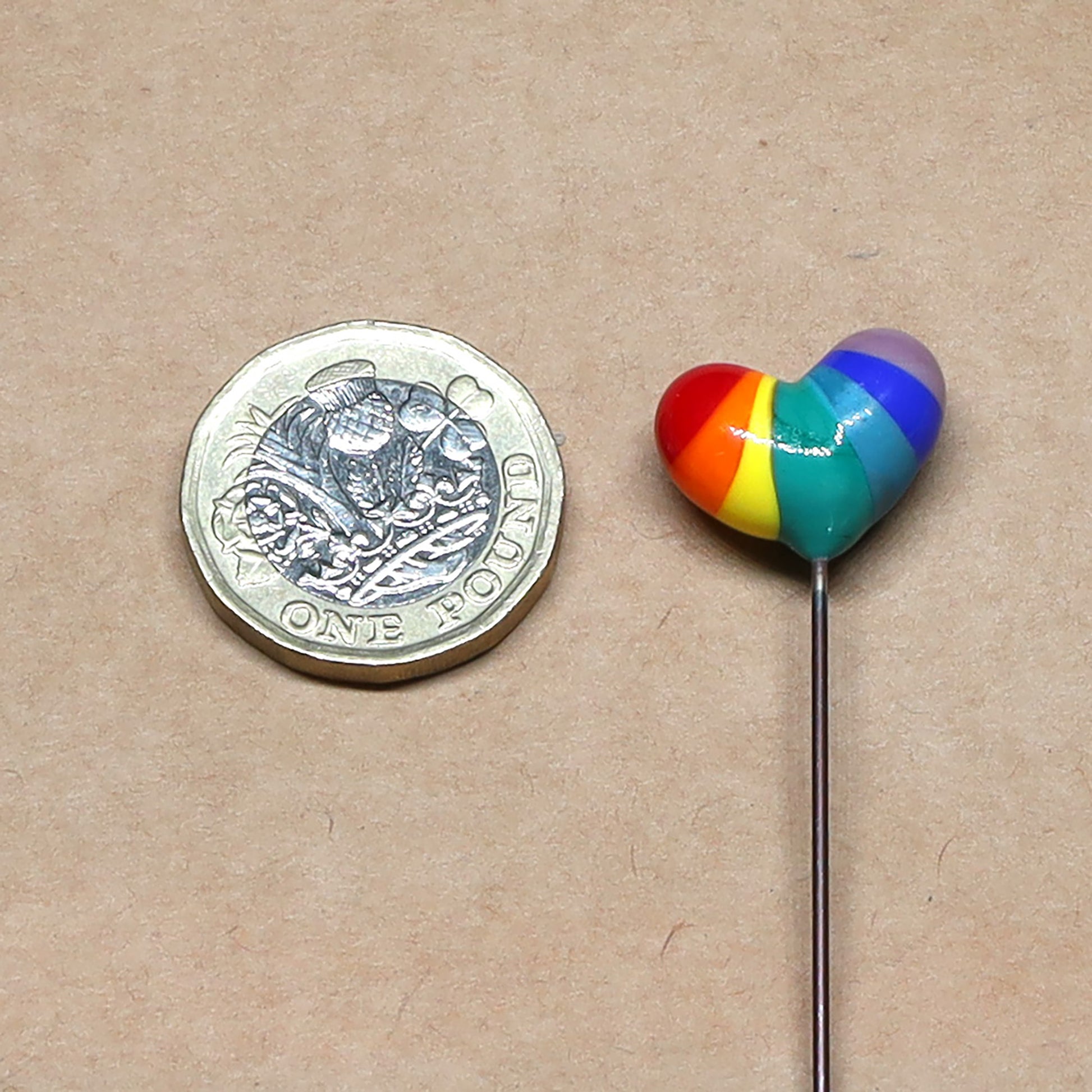 Size comparison showing a rainbow glass heart plant stake next to a one-pound coin on a neutral background, illustrating the small scale of the decoration