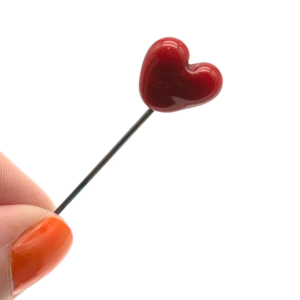 Tiny red glass heart on top of a pin shown held between a thumb and forefinger.