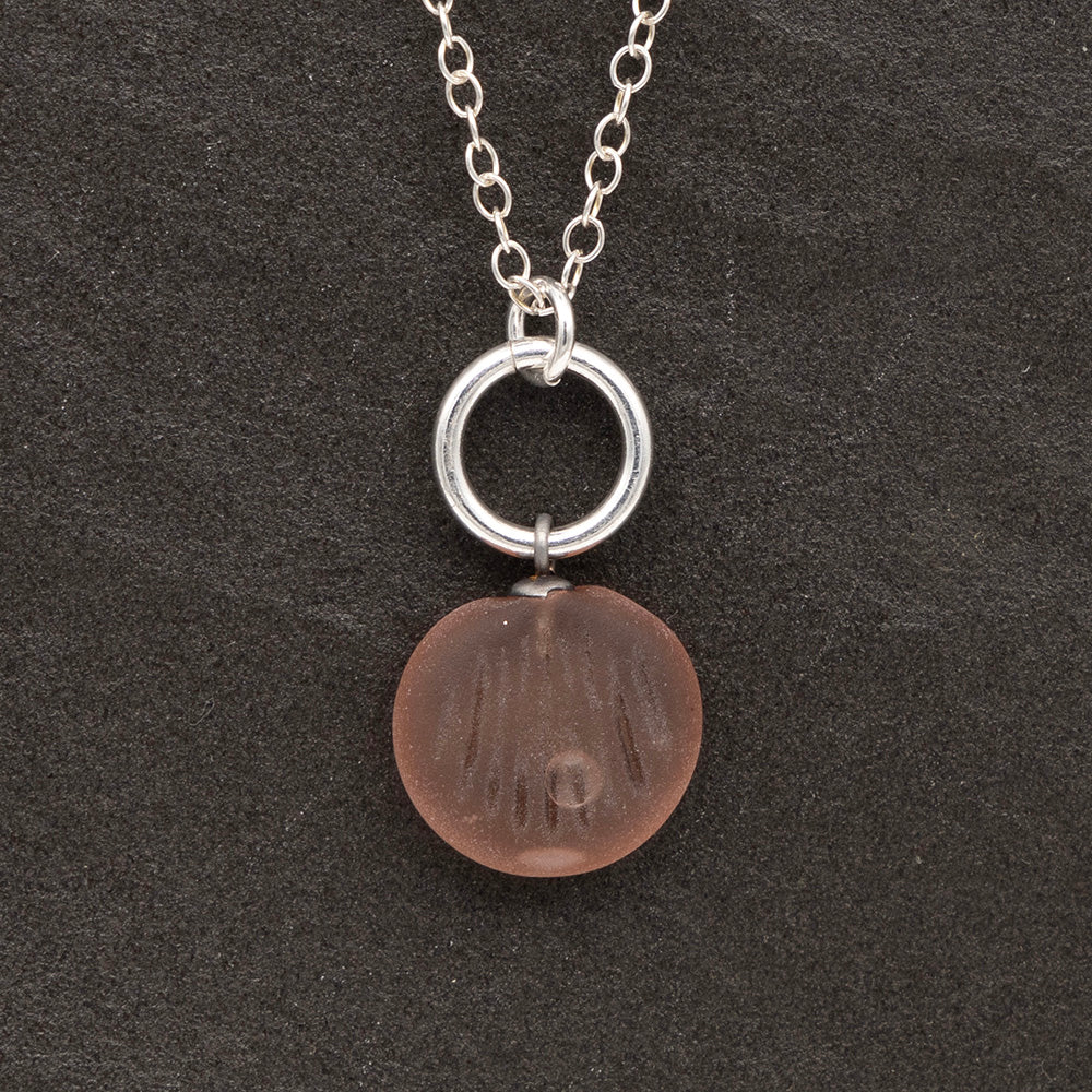 Close up of a pink frosted glass charm with shell imprint on a dark slate background. The charm is frosted, and spiral is shiny. The charm hangs on a large silver jump ring. The necklace is completed with a small silver jump ring and silver chain.