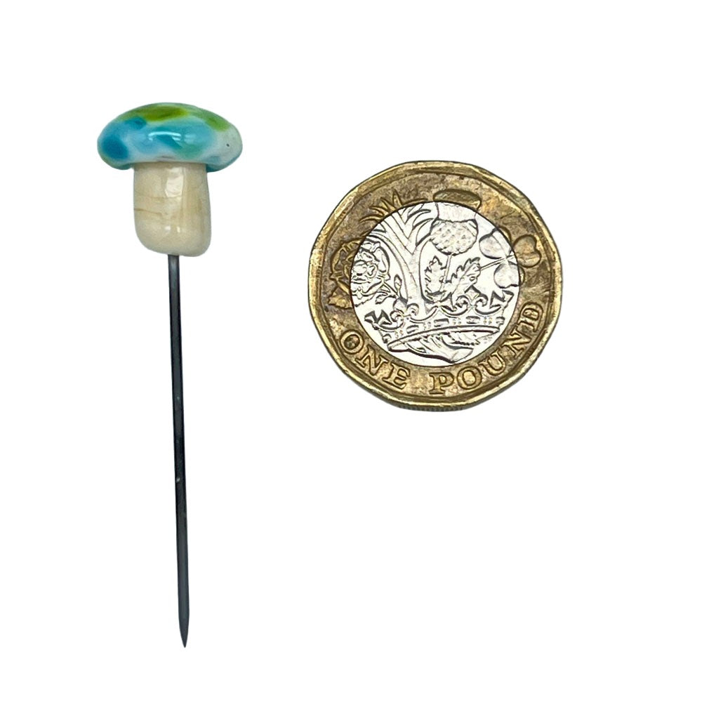 Speckled mushroom pin with green and blue cap next to a pound coin: A glass mushroom pin with a cap featuring turquoise, green and blue speckles, placed beside a one-pound coin to show size.