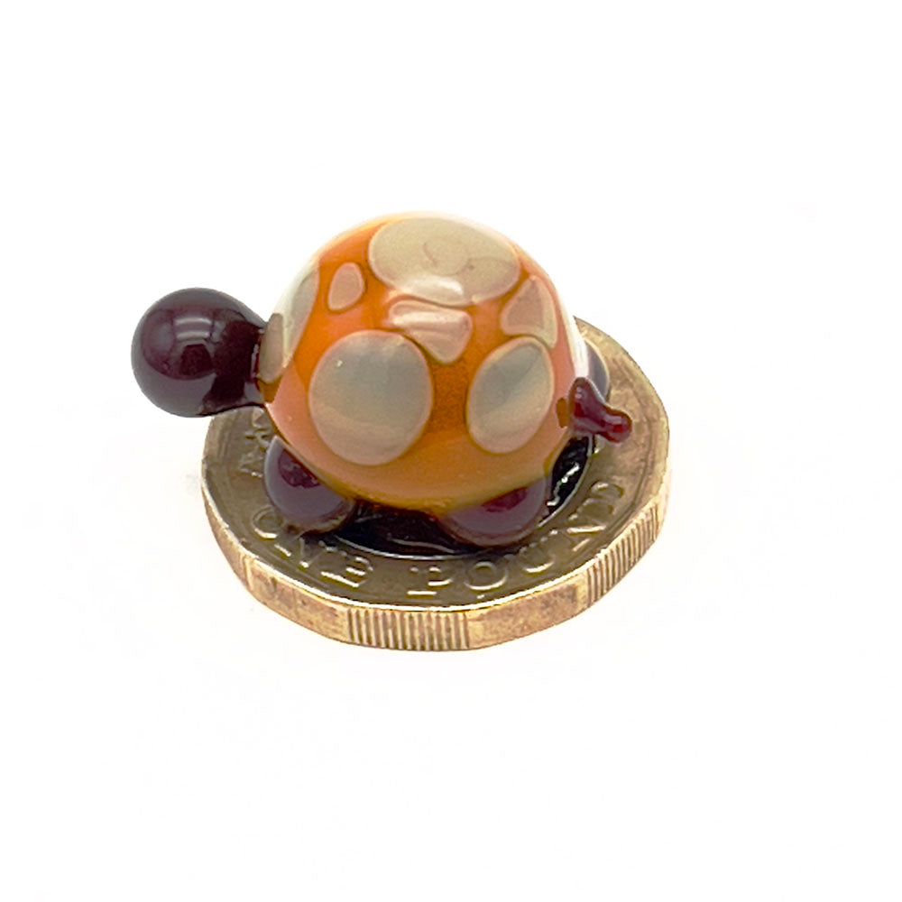 A yellow and grey glass tortoise with light grey spots placed on a one-pound coin for scale, viewed from the back.