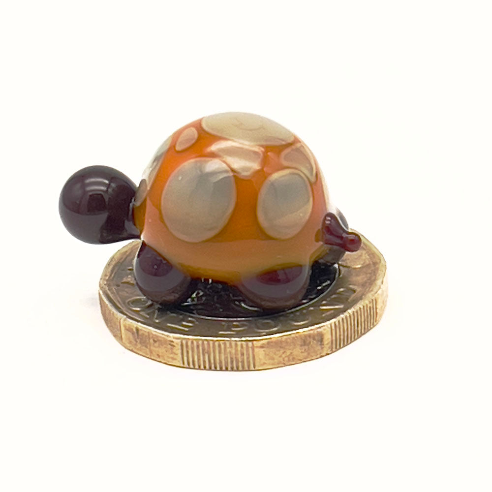 A yellow and grey glass tortoise with light grey spots placed on a one-pound coin for scale, viewed from the side.
