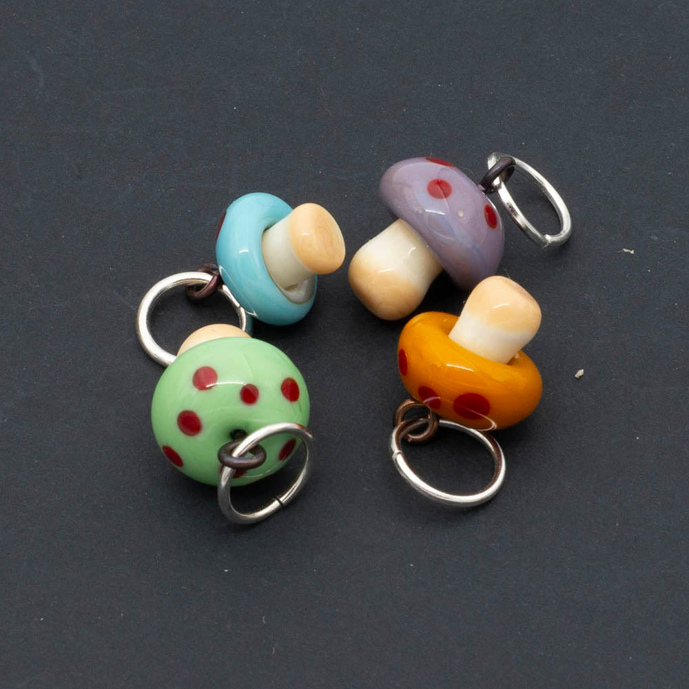 Four tiny glass mushroom stitch markers. The mushrooms have cream stems. Caps are green, blue, purple and yellow and all mushrooms have red dots, The mushrooms hand from jump rings.