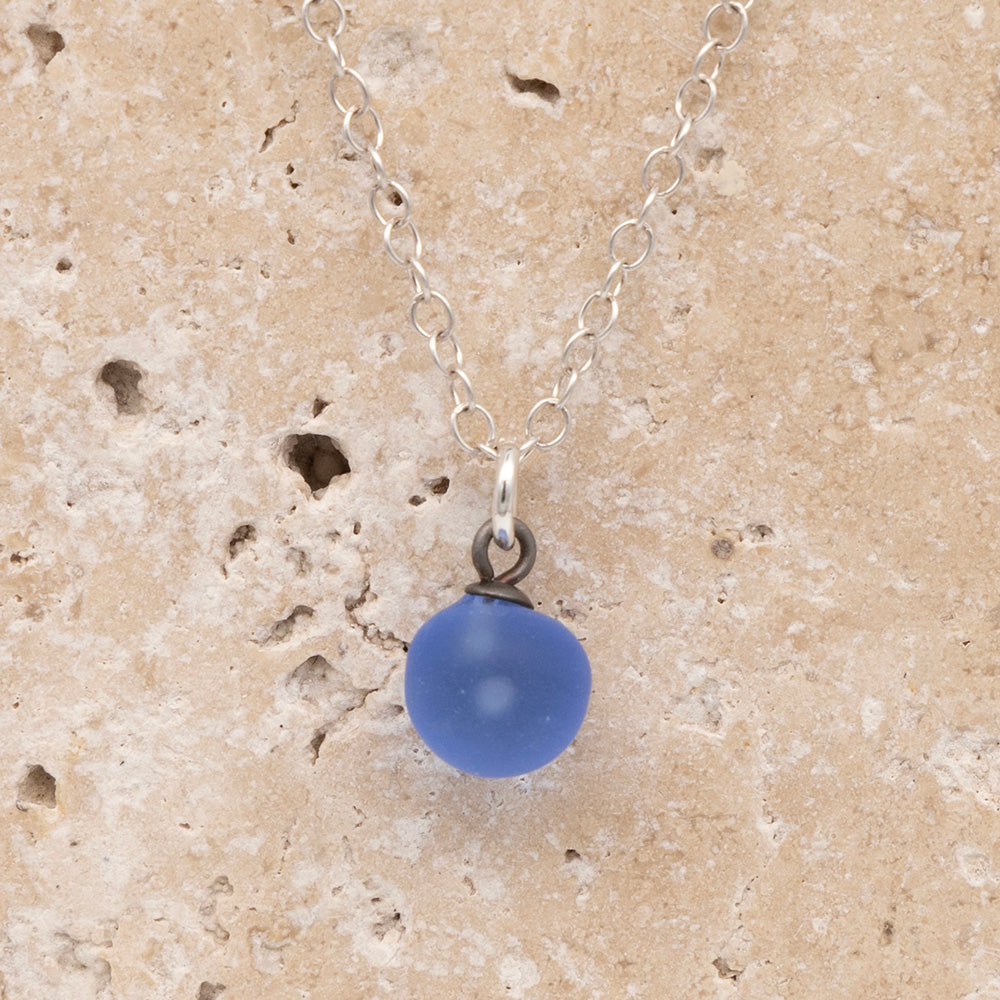 Close up of a blue frosted glass ball charm on a sandstone background. The charm is frosted. It hangs on a small silver jump ring. The necklace is completed with a silver chain.