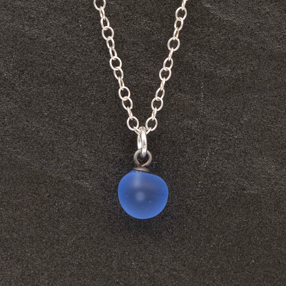 Close up of a blue frosted glass ball charm on a dark slate background. The charm is frosted. It hangs on a small silver jump ring. The necklace is completed with a silver chain.