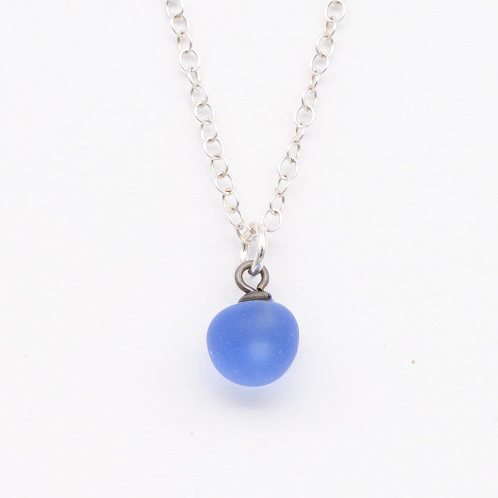 Close up of a blue frosted glass ball charm on a plain white background. The charm is frosted. It hangs on a small silver jump ring. The necklace is completed with a silver chain.