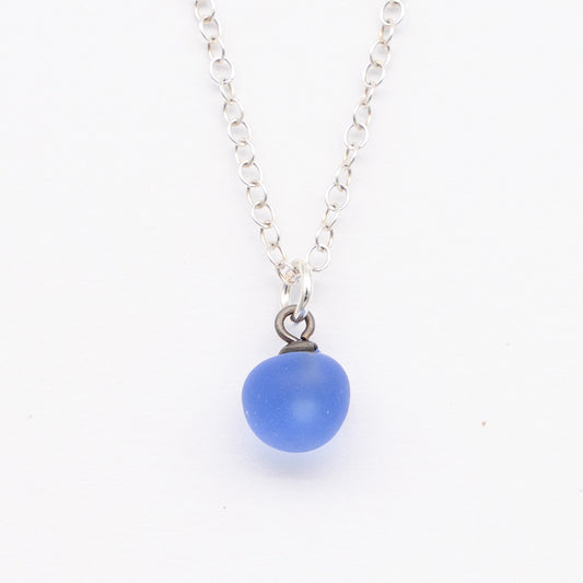 Close up of a blue frosted glass ball charm on a plain white background. The charm is frosted. It hangs on a small silver jump ring. The necklace is completed with a silver chain.