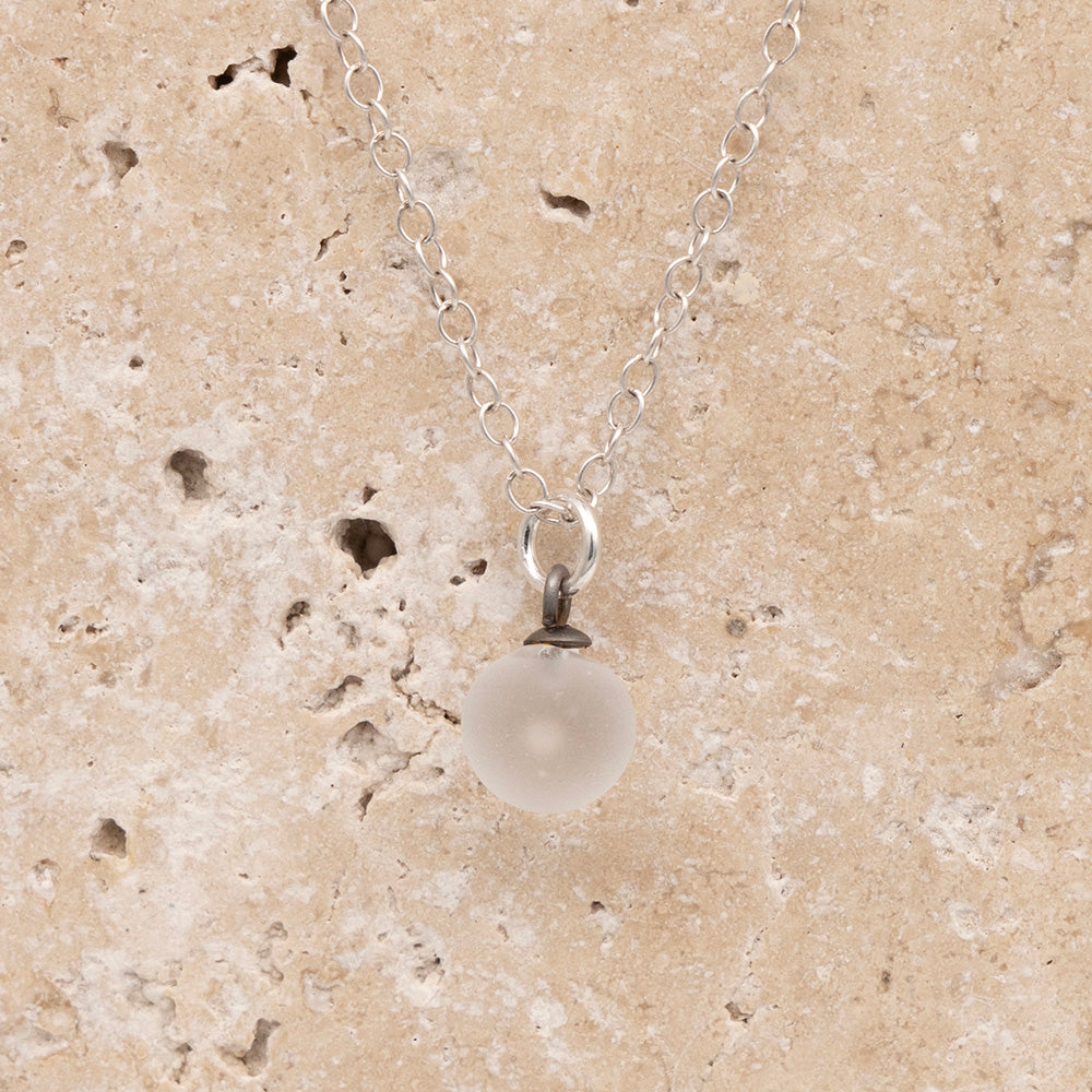 Close up of a clear frosted glass ball charm on a sandstone background. The charm is frosted. It hangs on a small silver jump ring. The necklace is completed with a silver chain.