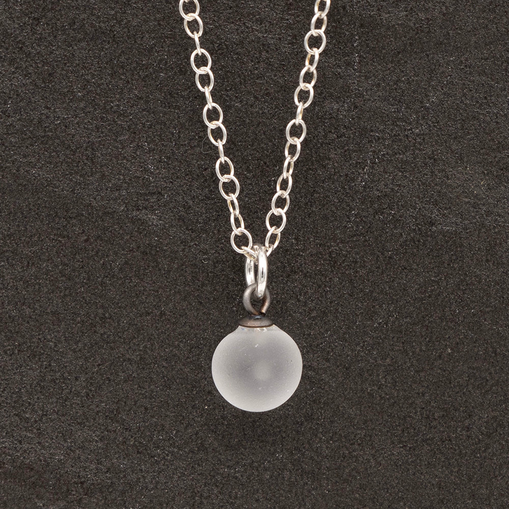 Close up of a clear frosted glass ball charm on a dark slate background. The charm is frosted. It hangs on a small silver jump ring. The necklace is completed with a silver chain.