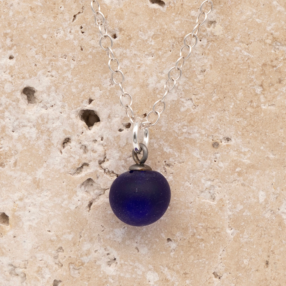 Close up of a dark blue frosted glass ball charm on a sandstone background. The charm is frosted. It hangs on a small silver jump ring. The necklace is completed with a silver chain.