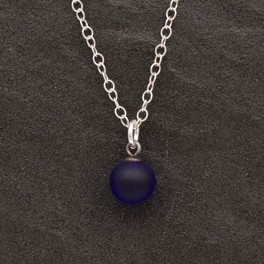 Close up of a dark blue frosted glass ball charm on a dark slate background. The charm is frosted. It hangs on a small silver jump ring. The necklace is completed with a silver chain.