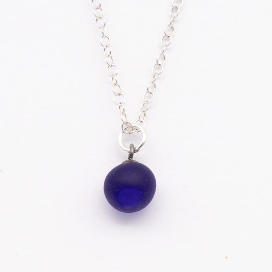 Close up of a dark blue frosted glass ball charm on a plain white background. The charm is frosted. It hangs on a small silver jump ring. The necklace is completed with a silver chain.