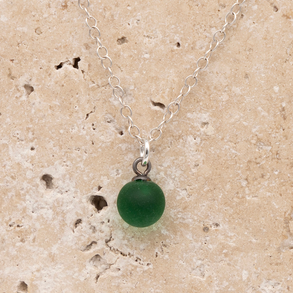 Close up of a green frosted glass ball charm on a sandstone background. The charm is frosted. It hangs on a small silver jump ring. The necklace is completed with a silver chain.