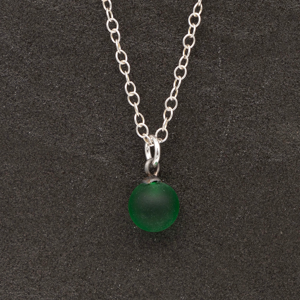 Close up of a green frosted glass ball charm on a dark slate background. The charm is frosted. It hangs on a small silver jump ring. The necklace is completed with a silver chain.