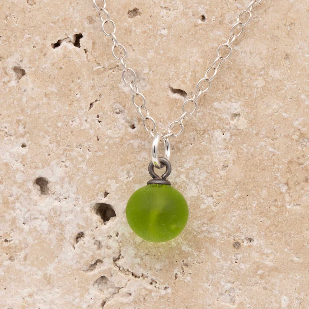 Close up of a lime green frosted glass ball charm on a sandstone background. The charm is frosted. It hangs on a small silver jump ring. The necklace is completed with a silver chain.