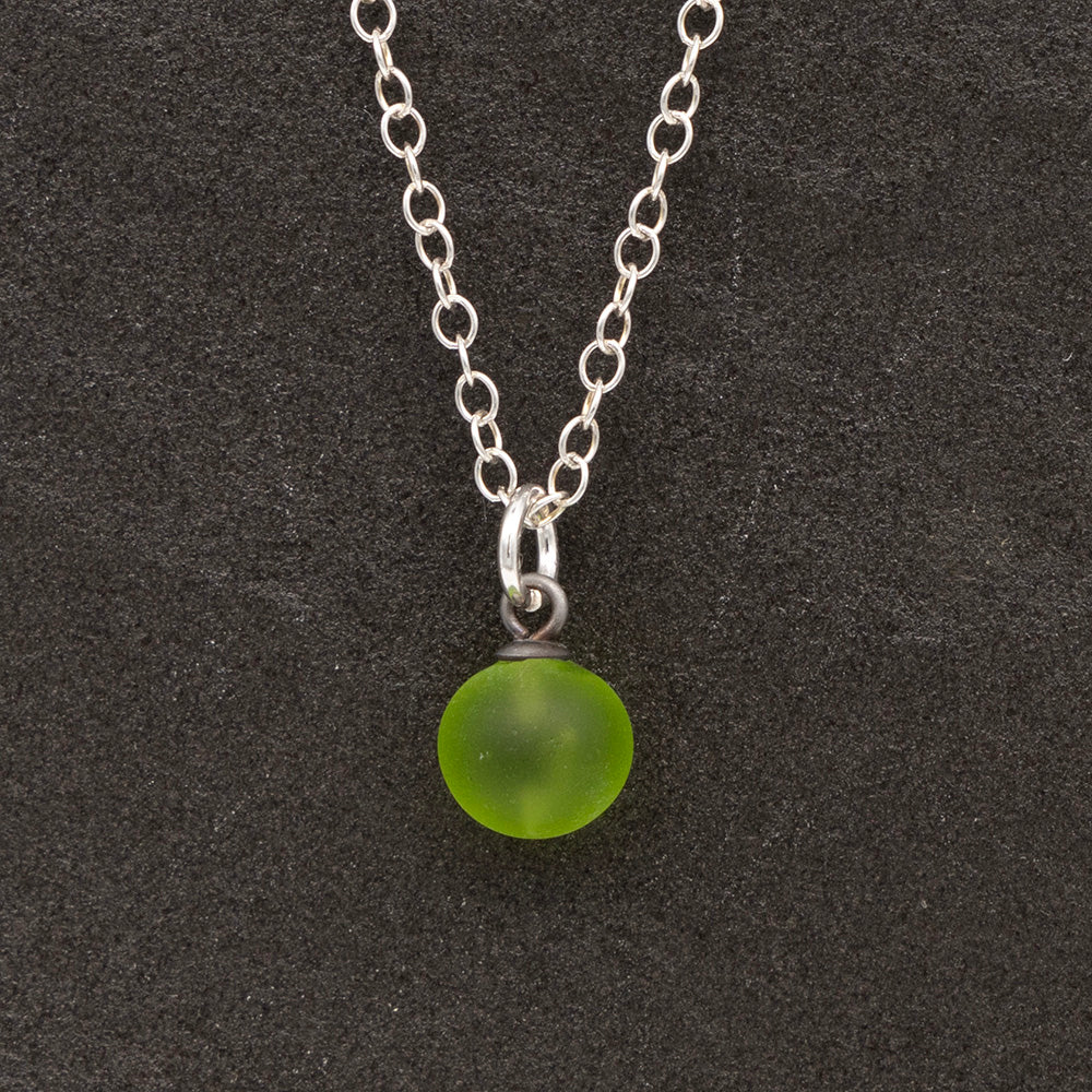 Close up of a lime green frosted glass ball charm on a dark slate background. The charm is frosted. It hangs on a small silver jump ring. The necklace is completed with a silver chain.